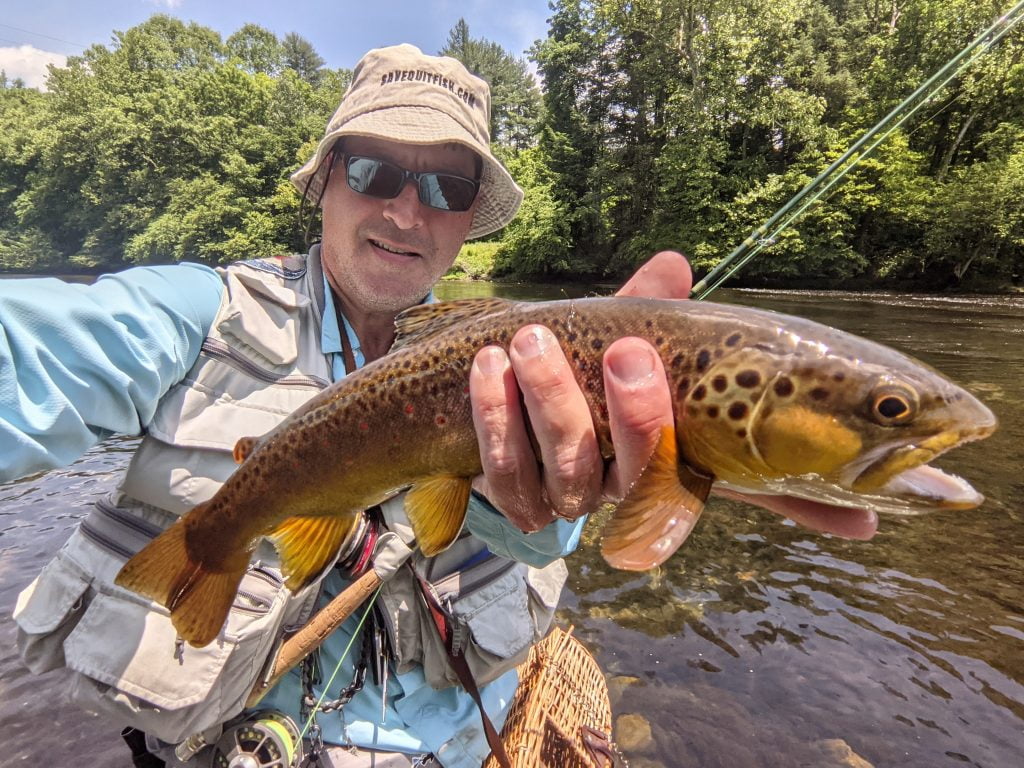 Fly fisherman smiles and holds wild brown trout in hand.
